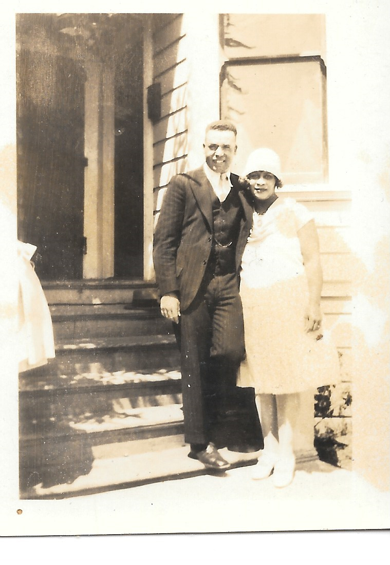A couple stands on the front stairs of a house in period clothing.